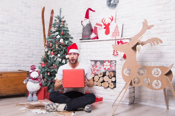 Happy holidays celebration. Man in santa hat sit on floor at fireplace. Christmas and new year presents preparation. Macho hold present box with surprised face at xmas tree. Boxing day concept.