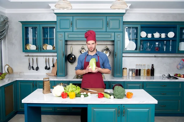 Guy in chef hat, apron hold chinese cabbage in kitchen — Stock Photo, Image