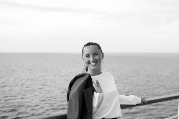 business woman or happy cute girl with smiling face in grey jacket, has red manicure on ship deck viewing sea, ocean water with sailing boat on cloudy sky background, traveling and tourism