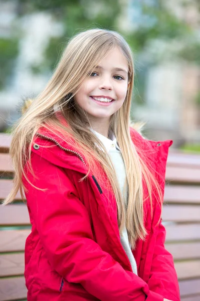 Kind mit blonden langen Haaren lächelt draußen. Glückliche Kindheit. Mädchen in rotem Mantel sitzen auf Bank im Park. Freizeit, Entspannung, Lebensstil. Kinder genießen den Herbsttag. — Stockfoto