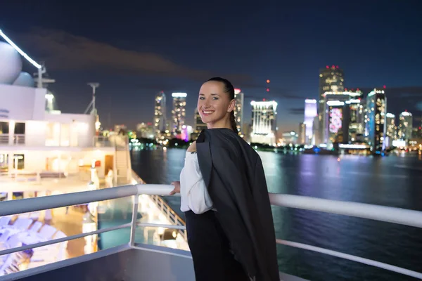 Mujer de negocios sonríe a bordo de un barco por la noche en Miami, EE.UU. Mujer sensual en chaqueta de traje en el horizonte de la ciudad. Moda, belleza, mira. Viajar por negocios. Lujuria, aventura, descubrimiento, viaje. — Foto de Stock