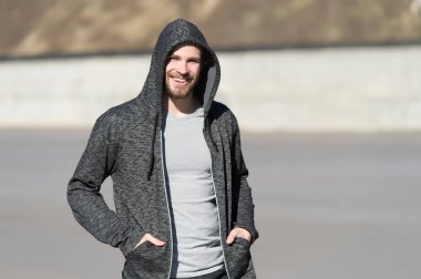 Kapüşonlu sakallı adam dışarıda güneşli bir şekilde gülümsüyor. Maço tişört içinde mutlu bir şekilde gülümsüyor, gündelik tarzda. Erkek modası, tarz, spor giyim. Aktif ve sağlıklı insanlar için yaşam tarzı, spor.