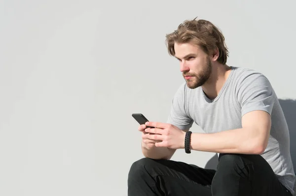 Uomo seduto con smartphone su parete grigia, nuova tecnologia. Macho con barba, capelli biondi in maglietta nella giornata di sole, moda. Nuova tecnologia, dispositivo mobile. Moda uomo con accessorio e stile, spazio copia — Foto Stock