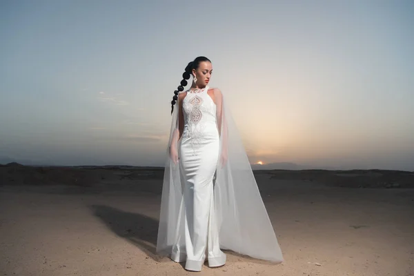 Bride in wedding gown on sunset sky. Woman in white dress in desert. Sensual woman with brunette hair. Fashion model in sand dunes. Beauty girl with glamour look and makeup — Stock Photo, Image
