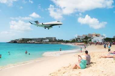 Philipsburg, Sint Maarten - February 13, 2016: jet flight land over maho beach. Plane low fly on cloudy blue sky. Airplane over people on sea. Beach vacation at Caribbean. Wanderlust, travel and trip clipart