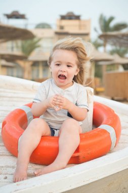 Güvenlik, kurtarma ve yardım kavramı. Mutlu çocuk güneşli günde yüzük şamandıra içinde oturmak. Lifebuoy tropik sahilde olan küçük çocuk. Eğlenceli açık sarı saçlı çocuk var. Yaz tatil ve eğlence aktivitesi