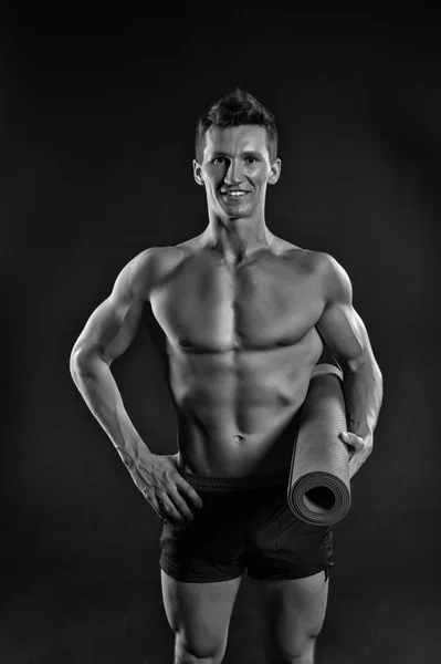 Atleta sonriendo con fuertes brazos en pantalones cortos. El hombre feliz sostiene la esterilla de yoga. Sonrisa deportiva con torso sexy y paquete de seis. Equipo de gimnasia para entrenamiento y entrenamiento. Deporte yoga y fitness, blanco y negro —  Fotos de Stock