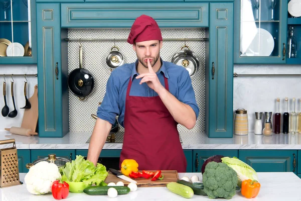 Mann kocht Geheimgericht in Küche. Koch mit schweigender Geste Finger an Tisch mit Gemüse. Zutaten zum Kochen von veganen Lebensmitteln. Vegetarisches Menü und gesunde Ernährung. Essenszubereitung und Kochrezepte — Stockfoto