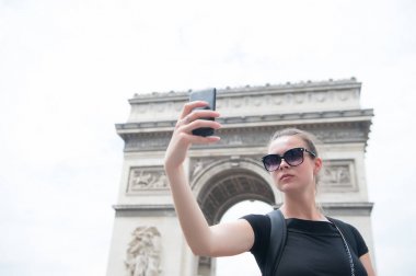 Paris, Fransa 'daki Arc de Triomphe' de bir kadın telefonla selfie çekiyor. Kemer Anıtı 'nda akıllı telefonu olan bir kadın. Fransa 'nın başkentinde tatil ve gezi. Moda görünümlü ve şehvetli bir kız.