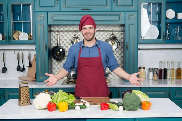 Mužský úsměv v kuchařském klobouku v kuchyni. Šťastná kuchařka u stolu. Zelenina a nástroje připravené k vaření. Příprava jídla a vaření recepty. Vegetariánské menu a zdravá strava — Stock fotografie