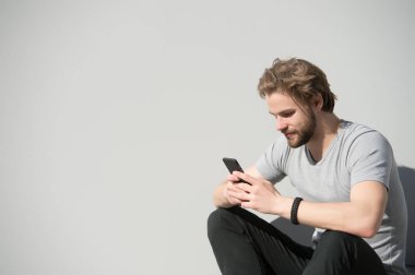 Sakallı adam gri duvarda cep telefonu kullanıyor. Güneşli açık havada akıllı telefonlu maço. Sakallı ve tişörtlü sarı saçlı bir adam mesaj attı. Yeni teknoloji ve modern hayat, boşluğu kopyala