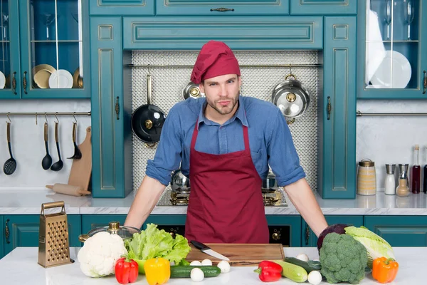 Schöner kaukasischer junger Mann in Schürze, der am Tisch mit Gemüse steht, zu Hause kocht und in einer Küche mit Holzoberfläche Mahlzeiten zubereitet, voller schickem Geschirr — Stockfoto
