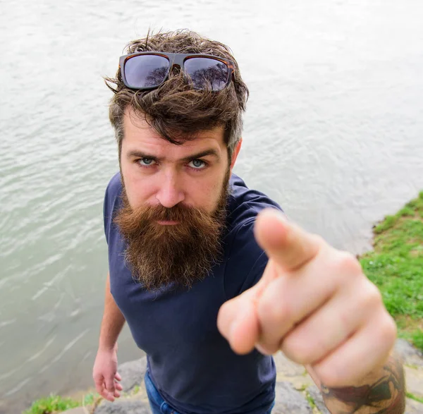 Uomo con barba e baffi con occhiali da sole, riva del fiume sullo sfondo. Hipster sul volto serio che punta con l'indice verso la fotocamera, sfocato. Indicazione e concetto di direzione — Foto Stock