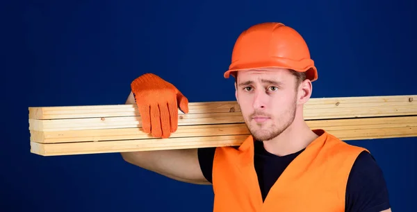 Snickare, möbelsnickare, stark builder på tankeväckande ansikte bär träbalk på axeln. Mannen i hjälm, hjälm och skyddshandskar innehar träbalk, blå bakgrund. Trä material koncept — Stockfoto