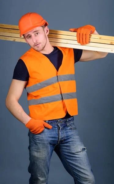 Carpintero, carpintero, obrero, constructor de cara segura lleva vigas de madera en el hombro. Hombre con casco, sombrero duro y guantes de protección sostiene la viga de madera, fondo gris. Concepto de trabajador resistente — Foto de Stock