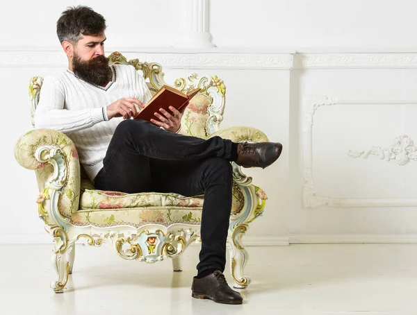 肘掛け椅子にひげと口ひげ、座っていると読んで、白の壁背景を持つ男。文学概念の目利き。鑑定・教授文学をお楽しみください。マッチョは本と余暇を費やしています。 — ストック写真