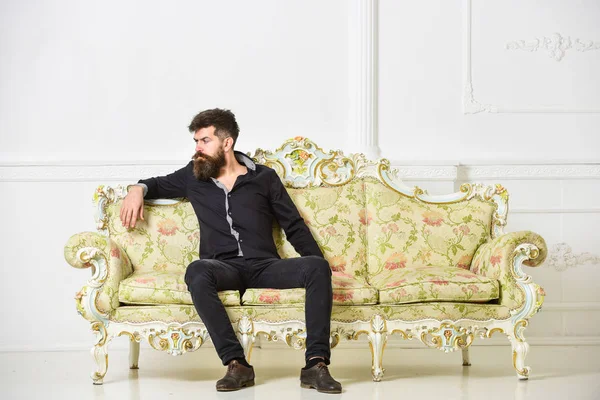 Hombre con barba y bigote pasa el ocio en la sala de estar de lujo. Hipster en cara pensativa se sienta solo. Concepto rico y solitario. Propietario de apartamento de lujo se sienta en el sofá, fondo blanco de la pared — Foto de Stock