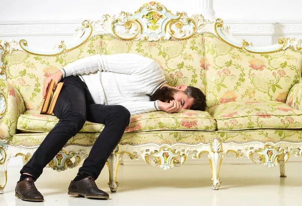 Macho sleep with book in hand. Boring literature concept. Man with beard and mustache lay on baroque style sofa, holds book, white wall background. Guy fall asleep while reading old boring book