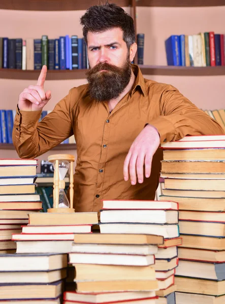 Homem no rosto confiante fica entre pilhas de livros, enquanto estuda na biblioteca, estantes de livros em segundo plano. Professor ou estudante com barba está à mesa com livros, desfocado. Conceito de bibliotecário — Fotografia de Stock