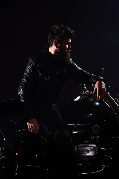 Homem com barba, motociclista de jaqueta de couro magra na moto do motor na escuridão, fundo preto. Brutalidade e conceito masculino. Macho, motociclista brutal em jaqueta de couro ficar perto de moto à noite — Fotografia de Stock