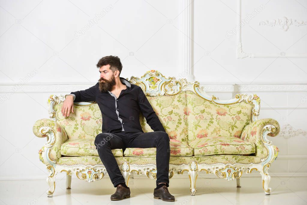 Man with beard and mustache spends leisure in luxury living room. Hipster on thoughtful face sits alone. Rich and lonely concept. Owner of luxury apartment sits on sofa, white wall background