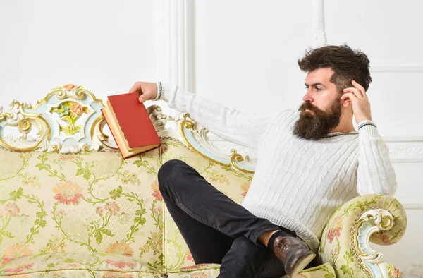 El hombre con barba y bigote pasa el ocio con el libro. Científico, profesor de cara estricta analizando literatura. Concepto de autoeducación. Profesor sentarse en el sofá y sostiene libro, fondo blanco de la pared —  Fotos de Stock