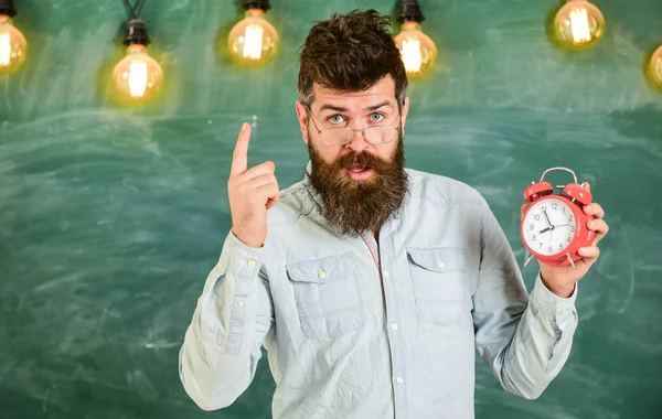 El profesor de anteojos tiene despertador. Concepto de disciplina. Hombre con barba y bigote en la cara estricta de pie en el aula. hipster barbudo sostiene el reloj, advertencia con el dedo índice, espacio de copia —  Fotos de Stock