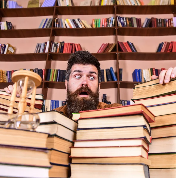 Muž na překvapený výraz mezi hromadami knih, při studiu v knihovně, regály na pozadí. Učitel nebo student s vousy sedí u stolu s knihami, rozostření. Bibliofil koncept — Stock fotografie