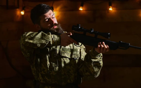 Férfi szakállal visel álcázás ruházati fából készült belső háttérben. Emlékek rabjai koncepció. Macho-a szigorú arcát gamekeepers házában. Hunter, brutális csípő fegyverrel a kezében készen áll a vadászat — Stock Fotó