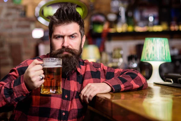 Hipster with beard holds glass with beer, raising up, cheers. Cheers concept. Guy spend leisure in bar, defocused background. Man on calm or serious face sit in bar or pub near bar counter — Stock Photo, Image