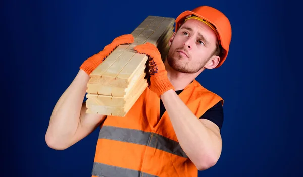 Carpenter, woodworker, strong builder on thoughtful face carries wooden beam on shoulder. Wooden materials concept. Man in helmet, hard hat and protective gloves holds wooden beam, blue background — Stock Photo, Image