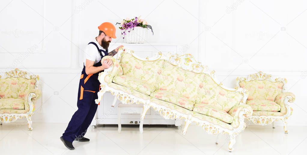 Loader moves sofa, couch. Courier delivers furniture in case of move out, relocation. Delivery service concept. Man with beard, worker in overalls and helmet lifts up sofa, white background