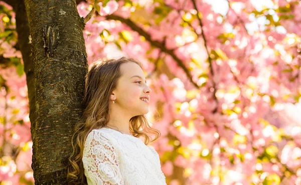 Dziewczyna z długimi włosami odkryty, wiśni lub sakura na tle. Słodkie dzieci cieszyć się przyrodą na dzień wiosny. Dziewczyna na uśmiechniętą twarz, stojący w pobliżu sakura, oprzeć się na pniu drzewa. Naturalne piękno pojęcie — Zdjęcie stockowe