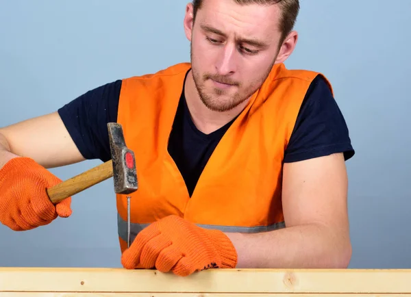 Carpenter, woodworker on concentrated face hammering nail into wooden board. Man, handyman in bright vest and protective gloves handcrafting, light blue background. Handcrafting concept