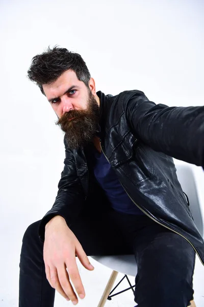 Man with beard and mustache on strict face looks at camera. Macho wears leather jacket, white background. Menswear and fashion concept. Hipster looks serious while sitting on chair in stylish outfit