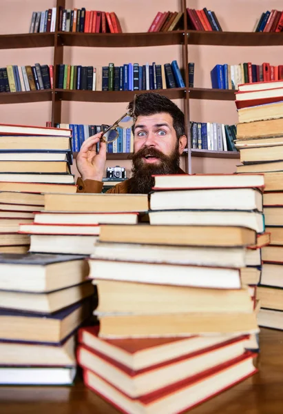 Δάσκαλος ή μαθητής με γενειάδα φοράει γυαλιά, κάθεται στο τραπέζι με τα βιβλία, defocused. Έννοια της επιστημονικής έρευνας. Ο άνθρωπος στο πρόσωπο έκπληξη μεταξύ σωρούς από βιβλία στη βιβλιοθήκη, ράφια σε φόντο — Φωτογραφία Αρχείου