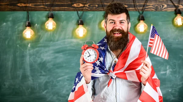 Έννοια του αμερικανικού εκπαιδευτικού συστήματος. Αμερικανός δάσκαλος με αμερικανικές σημαίες κατέχει ξυπνητήρι. Άνδρας με γενειάδα για χαρούμενο πρόσωπο κατέχει σημαία των ΗΠΑ και ρολόι στην τάξη, φόντου, Μαυροπίνακας, αντίγραφο χώρου — Φωτογραφία Αρχείου