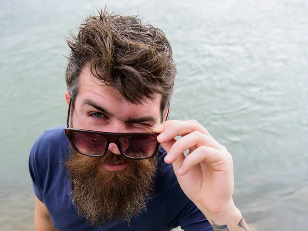 Eye protection concept. Man with beard and mustache wears sunglasses, water surface on background. Hipster on winking face peeking out of stylish sunglasses. Guy looks cool with stylish sunglasses