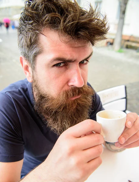 Hipster on serious strict face drinking coffee outdoor. Coffee break concept. Man with beard and mustache holds cup of coffee while relaxing at cafe terrace. Guy having rest with espresso coffee