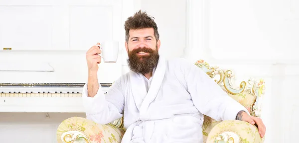 Веселый парень в белом халате наслаждается утренним кофе. Бородатый мужчина сидит в красивом антикварном кресле. Энергетическое утро дома — стоковое фото