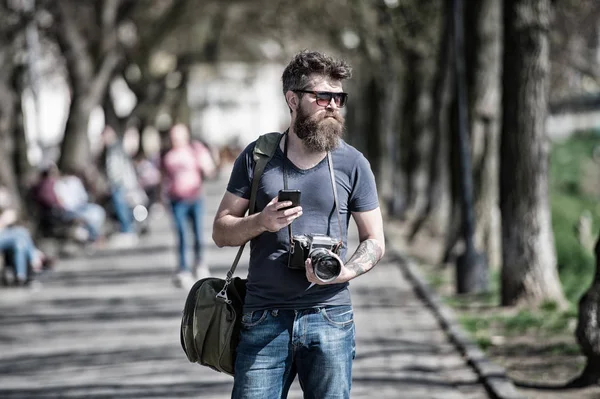 El hombre barbudo lleva la bolsa en el hombro y sostiene la cámara. Concepto de fotógrafo. Hombre con barba larga y elegante usando un teléfono inteligente mientras camina. Hombre con barba en rostro confiado, fondo urbano, desenfocado —  Fotos de Stock