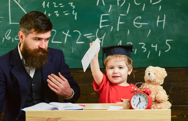 Little boy shows his copybook with paintings. Kid happy studies individually with father, at home. Individual schooling concept. Teacher and pupil in mortarboard, chalkboard on background