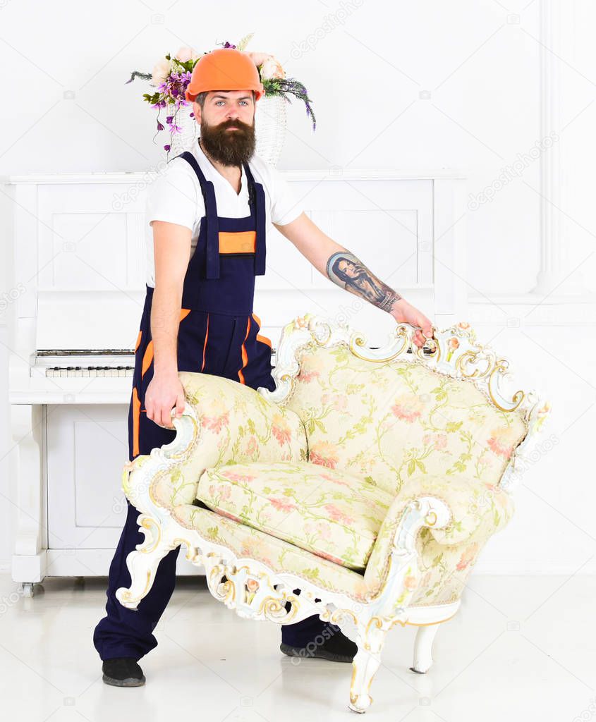 Delivery service concept. Courier delivers furniture in case of move out, relocation. Loader carries armchair. Man with beard, worker in overalls and helmet lifts up armchair, white background