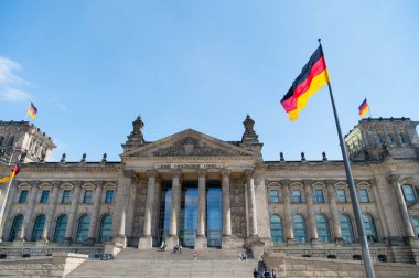 Alman bayrakları, Alman Parlamentosu 'nun (Deutscher Bundestag) ünlü binasında, mavi gökyüzü ve bulutlarla güneşli bir günde, Almanya' nın orta Berlin Mitte bölgesinde rüzgarda sallanıyor.