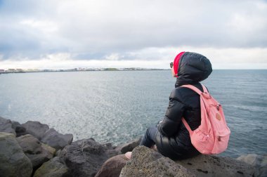 Reykjavik, İzlanda'taş deniz kıyısında kadın backpacker sit. Kadın Deniz Manzaralı bulutlu gökyüzü üzerinde keyfini çıkarın. Kız sırt çantası ile seyahat. Seyahat ve Yolculuk tutkusu ile tatil