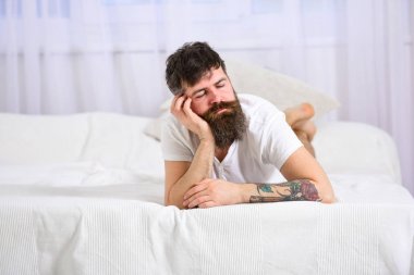 Guy on calm face sleeping on edge of bed. Exhausted concept. Man laying on bed, falling asleep while lean on hand, white curtains on background. Macho with beard sleeping, relaxing, having nap, rest clipart