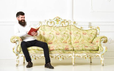 Sakal ve bıyık oturur Barok tarzı kanepe, tutar kitap, beyaz duvar arka plan üzerinde olan adam. Maço yüz kitap okuma konusunda tutkulu gülümseyen üzerinde. Adam zevk ile eski kitap okuma. Okuyucu kavramı