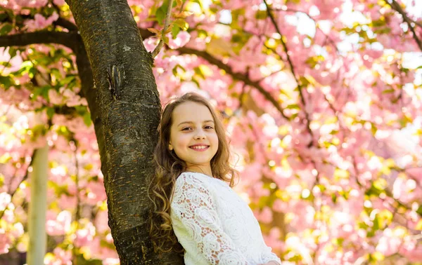 Meisje op het glimlachen gezicht permanent in de buurt van sakura, leunen op boomstam. Meisje met lang haar buiten, kersenbloesem of sakura op achtergrond. Schattig kind geniet van de natuur op de lente. Verenigen met natuur concept — Stockfoto