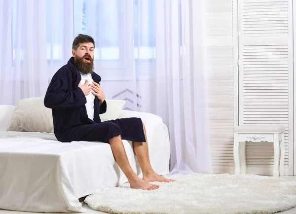 Guy on sleepy face yawning in morning. Awakening concept. Macho with beard and mustache sluggish yawning, relaxing after nap, rest. Man in robe sits on bed, white curtains on background