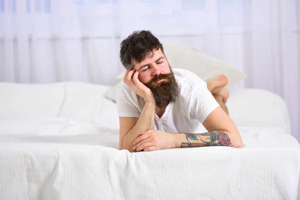 Un tipo con la cara tranquila durmiendo en el borde de la cama. Concepto agotado. Hombre acostado en la cama, durmiendo mientras se apoya en la mano, cortinas blancas en el fondo. Macho con barba durmiendo, relajándose, durmiendo la siesta, descansando —  Fotos de Stock